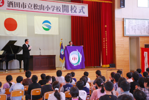 丸山市長（壇上左）から授与された校旗を、齋藤校長が児童たちに披露